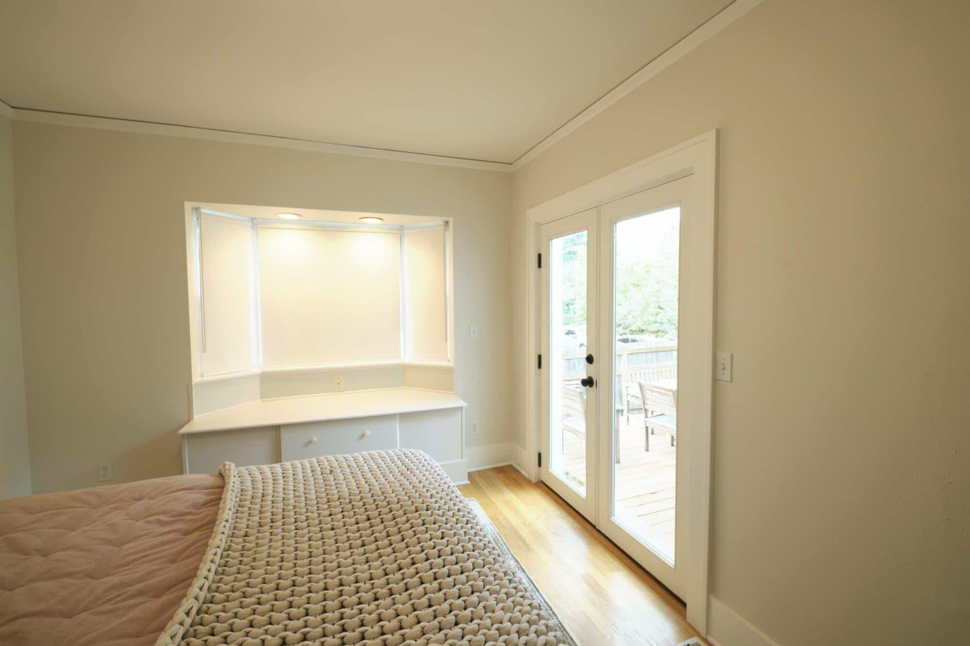 A cozy bedroom with a beige knitted blanket on the bed, a bay window with cabinets underneath, and French doors leading to an outdoor space. The room is well-lit with natural light.