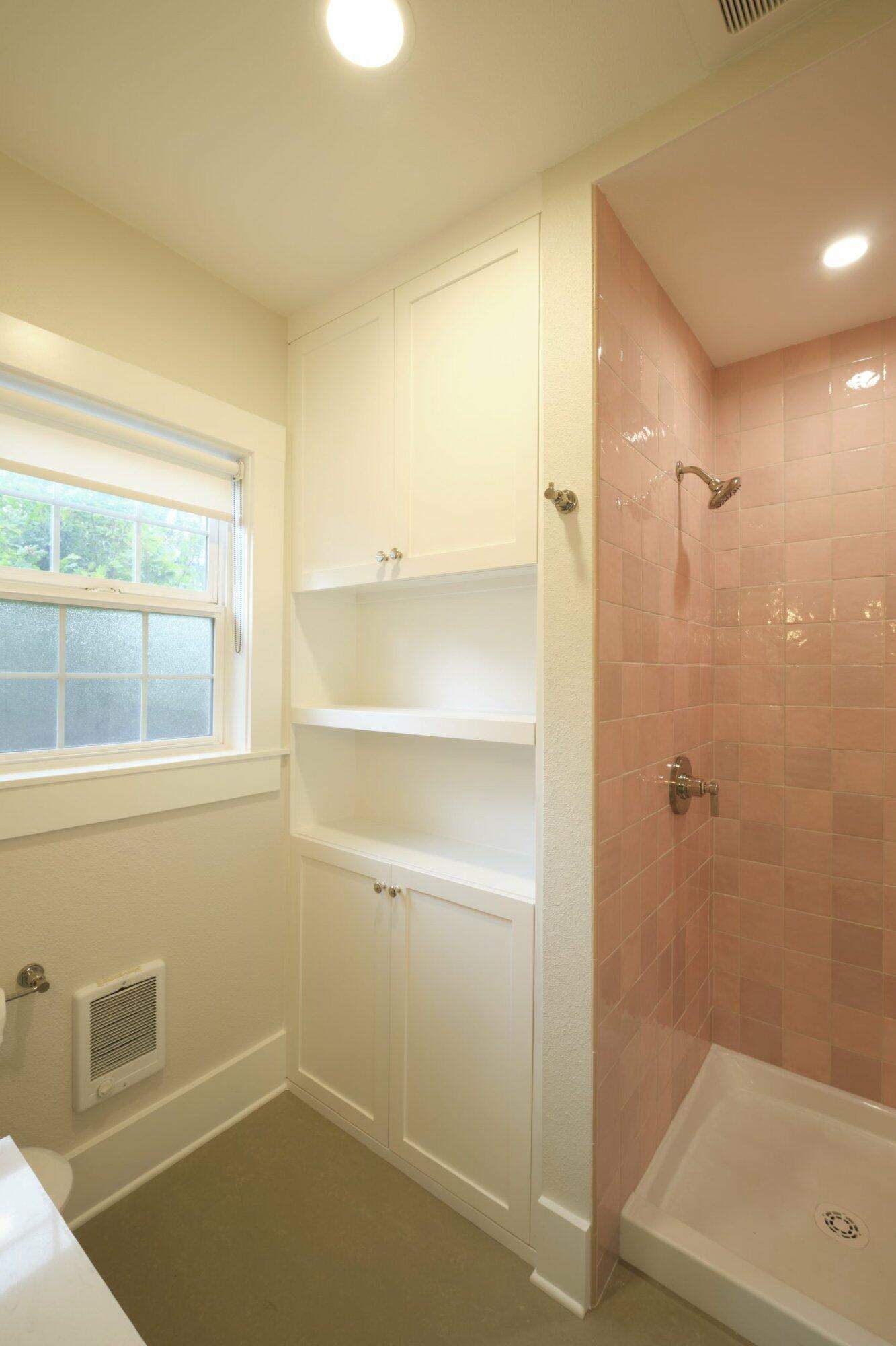 A bathroom with white shelves and cabinets, a window with blinds, and a shower area with pink tiled walls and a white shower base.