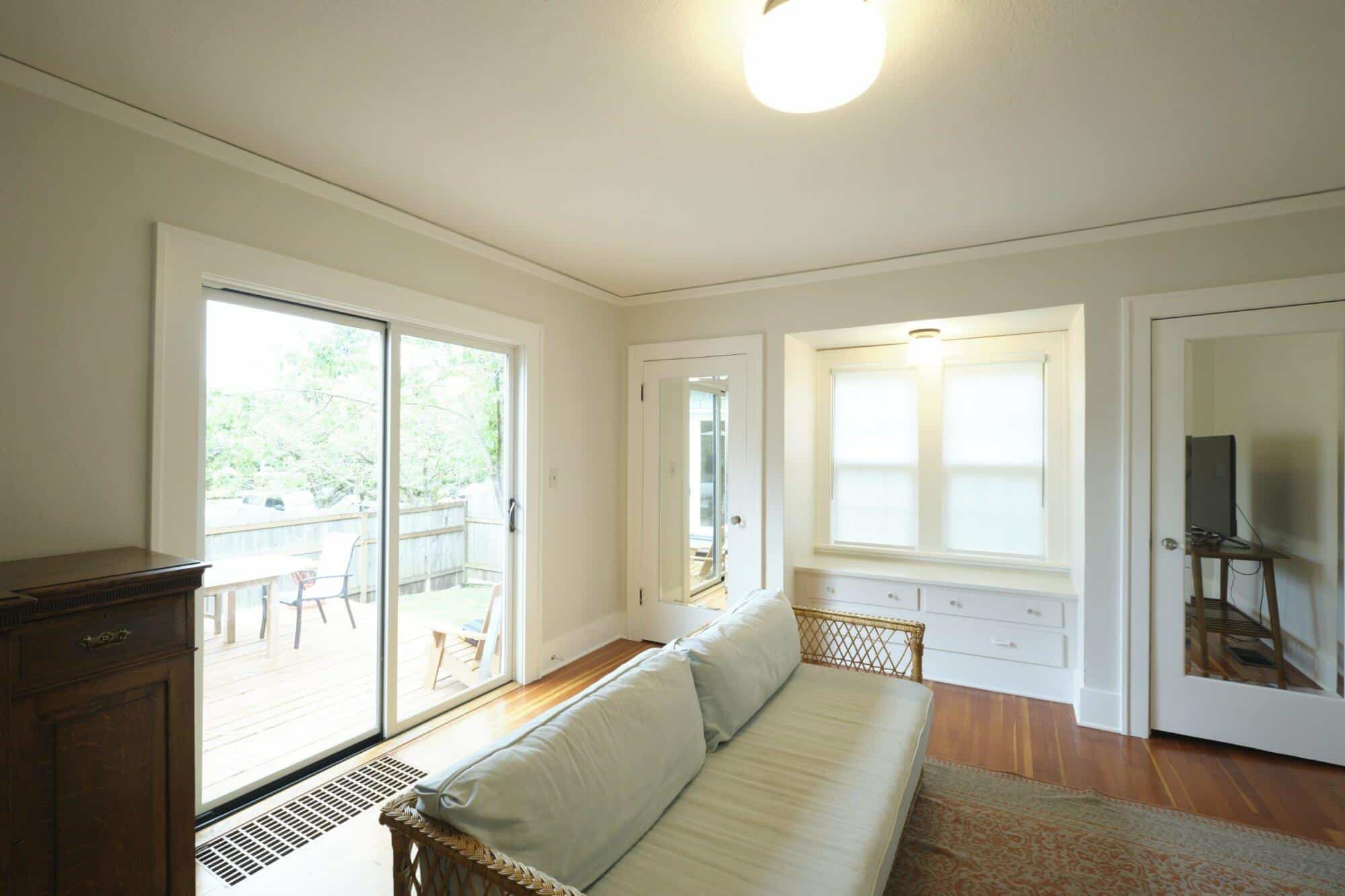 A bright living room featuring a vintage sofa, a glass door leading to a patio, wooden flooring, and a window with blinds. A television is partially visible on the right.