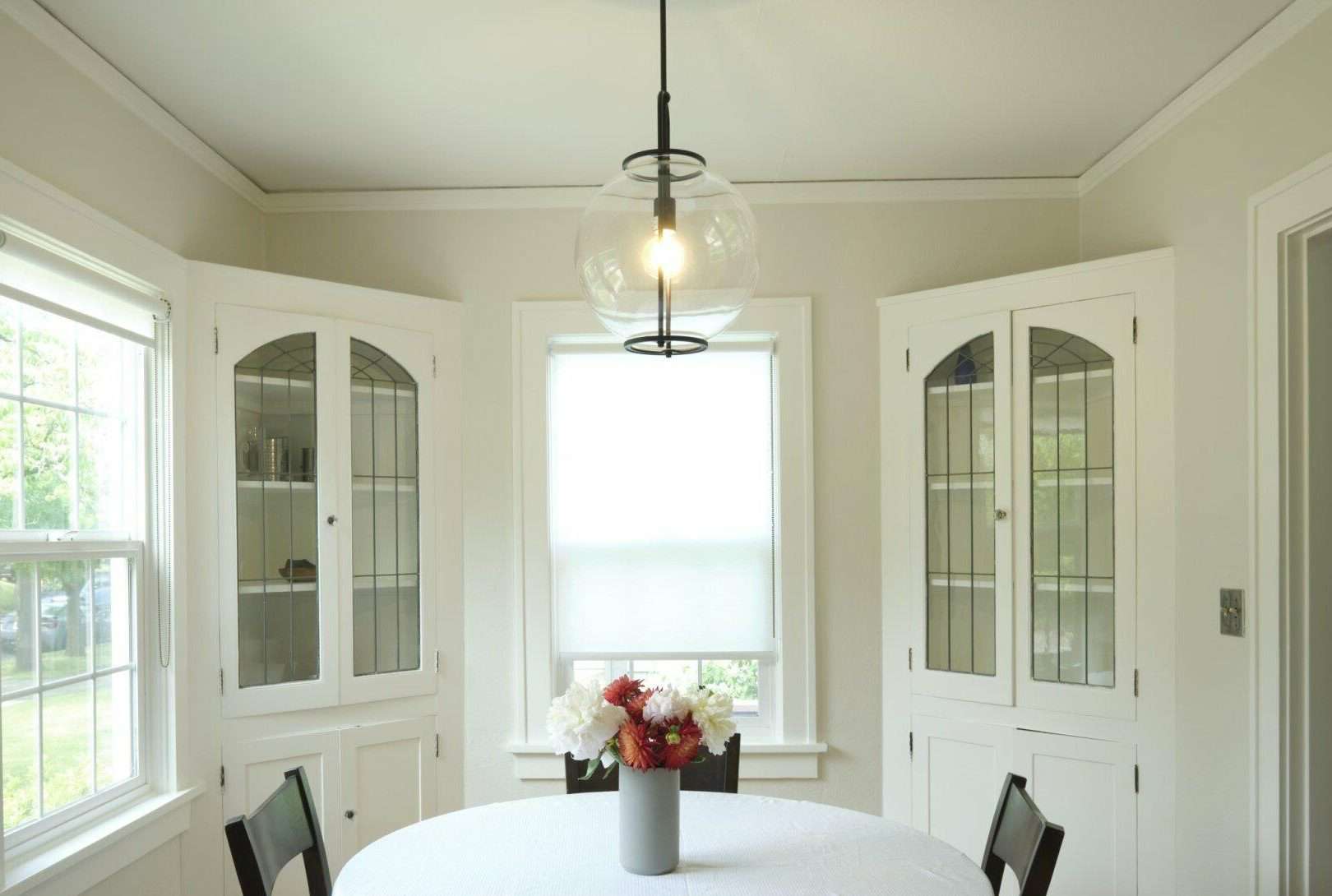 A dining area with a round table, a vase of flowers, two built-in corner cabinets with glass doors, and a single bulb pendant light hanging from the ceiling.