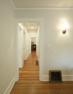 A hallway with wooden flooring, white walls, and a light fixture on the right wall. Multiple doors are along the sides, leading to other rooms, and the hallway leads to a room with a plant.
