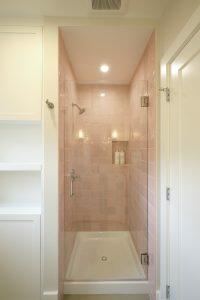 A small, enclosed shower with pink tiled walls, a clear glass door, and a white ceiling. Two bottles are visible on a ledge within the shower. Shelves are on the left, and a white door is on the right.