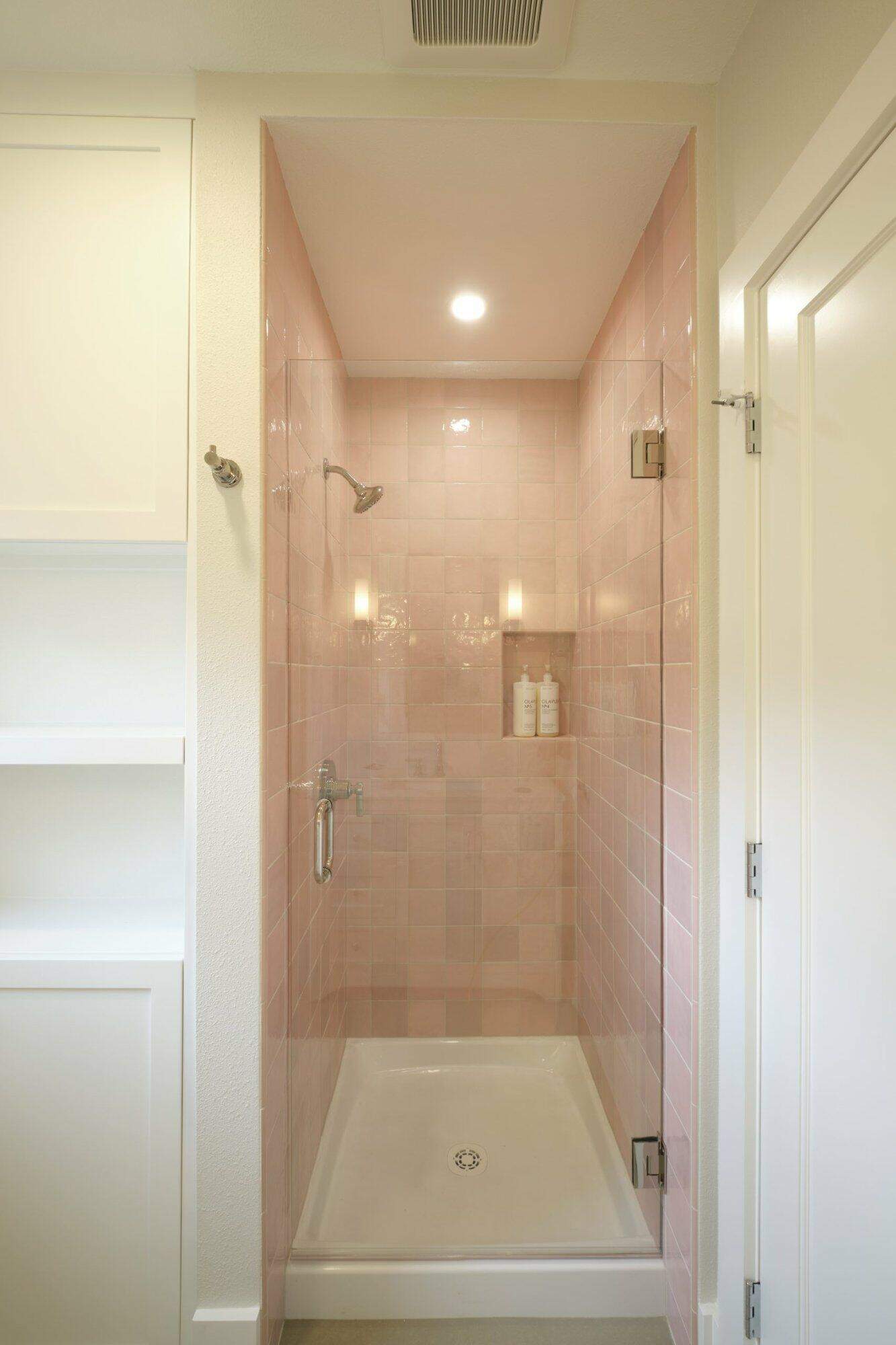 A small, enclosed shower with pink tiled walls, a clear glass door, and a white ceiling. Two bottles are visible on a ledge within the shower. Shelves are on the left, and a white door is on the right.
