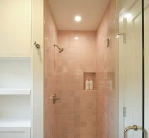 A small shower with pink square tiles, dual showerheads, a built-in shelf with bottles, a glass door, and an adjacent white shelving unit.