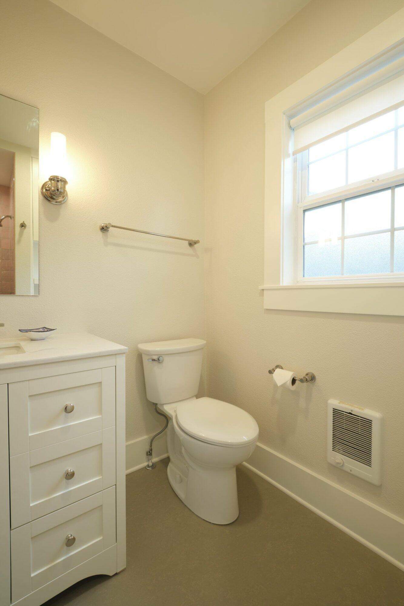 A small bathroom with a white toilet, a white vanity with drawers, a wall-mounted light above the mirror, a towel rack, a window with frosted glass, and a wall heater.