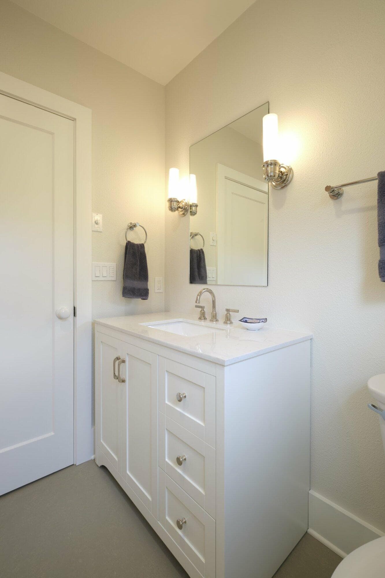 A bathroom with a white vanity featuring drawers and cabinet, a square mirror, two wall-mounted lights, a towel rack with a dark towel, and a closed door on the left.
