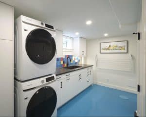 Laundry room with stacked washer and dryer, blue floor, colorful backsplash, white cabinets, a sink, and wall-mounted drying rack. Decor includes a framed picture.
