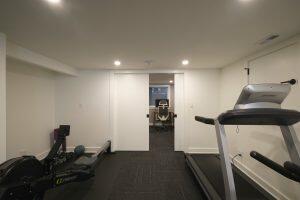 A home gym with exercise equipment including a treadmill and rowing machine, adjacent to a home office featuring a desk and office chair.