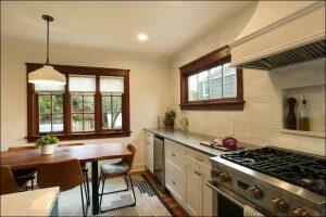 A modern kitchen with a wooden dining table, brown chairs, white cabinets, a black gas stove, and a window with wooden trim. A small plant and various kitchen items are on the counter. A modern kitchen with a wooden dining table, brown chairs, white cabinets, a black gas stove, and a window with wooden trim. A small plant and various kitchen items are on the counter.