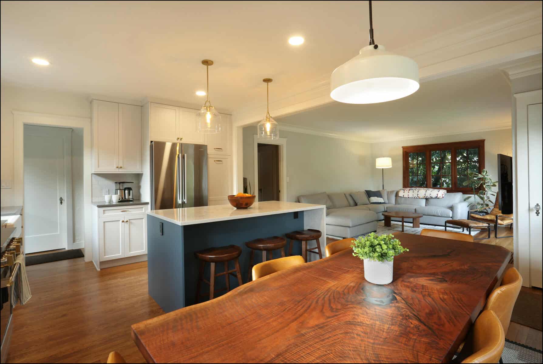 Modern kitchen with a central island featuring three stools, stainless steel appliances, and a wooden dining table with a plant centerpiece. The adjoining living area has a large couch and windows. Modern kitchen with a central island featuring three stools, stainless steel appliances, and a wooden dining table with a plant centerpiece. The adjoining living area has a large couch and windows.