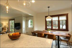 A modern dining area with a wooden table and six chairs, adjacent to a living room with a wall-mounted TV. A bowl of fruit sits on a marble countertop in the foreground. A modern dining area with a wooden table and six chairs, adjacent to a living room with a wall-mounted TV. A bowl of fruit sits on a marble countertop in the foreground.