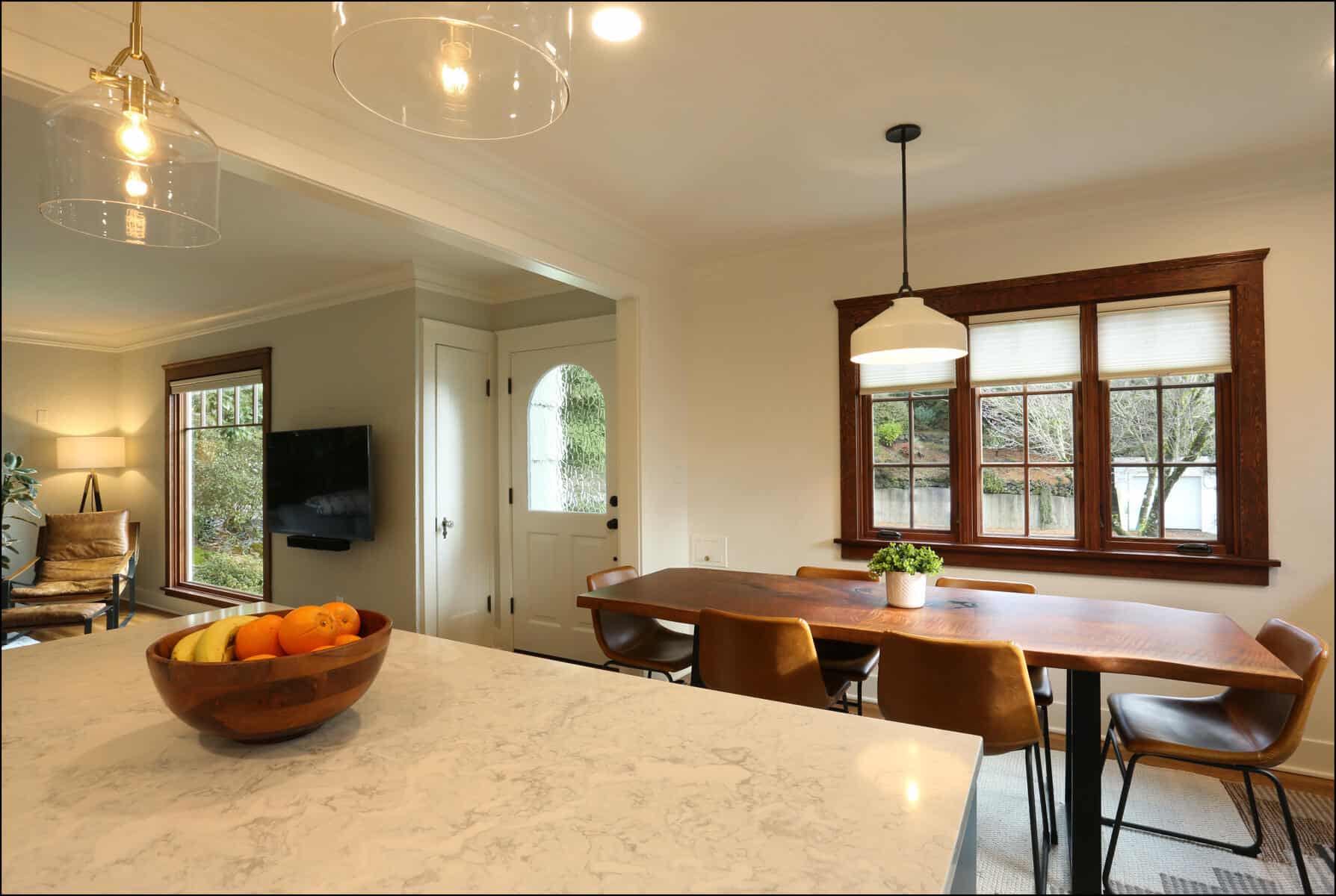 A modern dining area with a wooden table and six chairs, adjacent to a living room with a wall-mounted TV. A bowl of fruit sits on a marble countertop in the foreground. A modern dining area with a wooden table and six chairs, adjacent to a living room with a wall-mounted TV. A bowl of fruit sits on a marble countertop in the foreground.