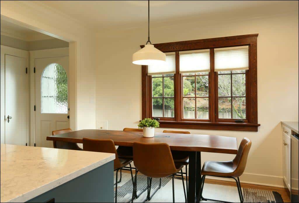 A dining area with a wooden table, four brown chairs, a potted plant centerpiece, and a window with a view of greenery. A white pendant light hangs above the table, and a door is visible in the background. A dining area with a wooden table, four brown chairs, a potted plant centerpiece, and a window with a view of greenery. A white pendant light hangs above the table, and a door is visible in the background.