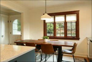 A dining area with a wooden table, four brown chairs, a potted plant centerpiece, and a window with a view of greenery. A white pendant light hangs above the table, and a door is visible in the background. A dining area with a wooden table, four brown chairs, a potted plant centerpiece, and a window with a view of greenery. A white pendant light hangs above the table, and a door is visible in the background.