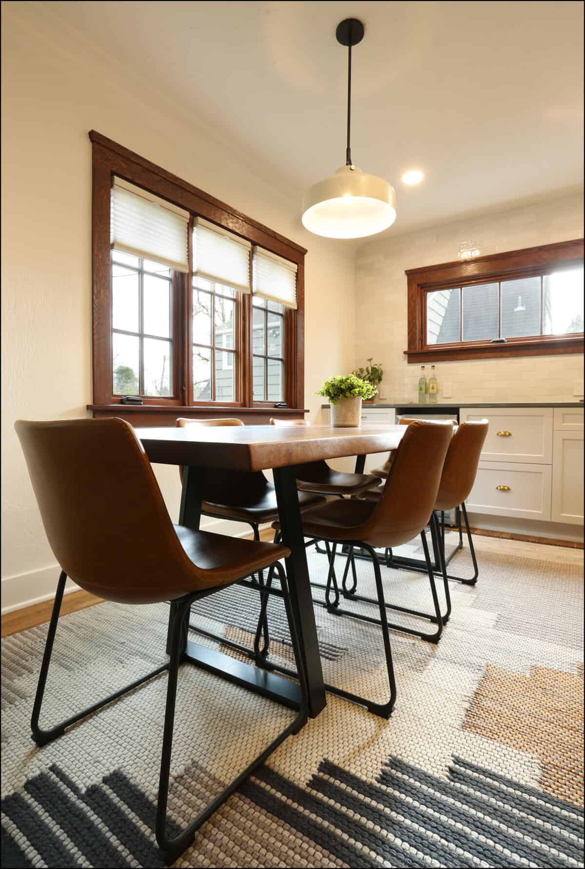 A dining room with a wooden table, four brown leather chairs, a ceiling pendant light, a potted plant on the table, and a window with white blinds in the background. A dining room with a wooden table, four brown leather chairs, a ceiling pendant light, a potted plant on the table, and a window with white blinds in the background.