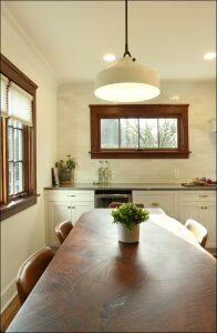 A kitchen with a wooden table, leather chairs, a potted plant, and a large pendant light. There is a counter with bottles and kitchenware, and a window with wooden trim above the counter. A kitchen with a wooden table, leather chairs, a potted plant, and a large pendant light. There is a counter with bottles and kitchenware, and a window with wooden trim above the counter.