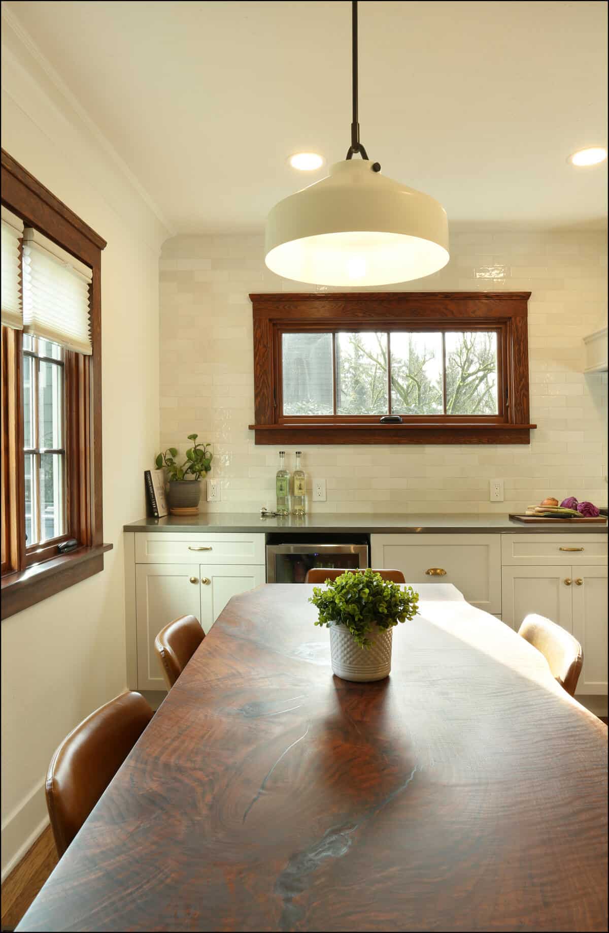 A kitchen with a wooden table, leather chairs, a potted plant, and a large pendant light. There is a counter with bottles and kitchenware, and a window with wooden trim above the counter. A kitchen with a wooden table, leather chairs, a potted plant, and a large pendant light. There is a counter with bottles and kitchenware, and a window with wooden trim above the counter.