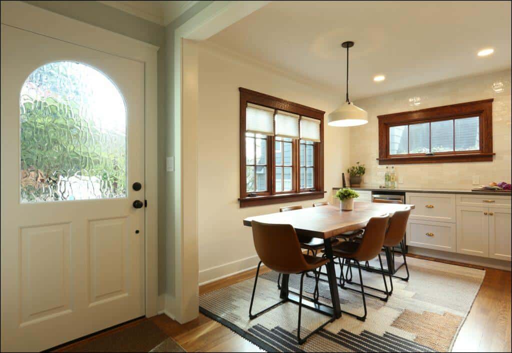 A well-lit dining area with a wooden table, four brown chairs, a hanging light fixture, and windows. The entry door has a textured glass panel. A well-lit dining area with a wooden table, four brown chairs, a hanging light fixture, and windows. The entry door has a textured glass panel.