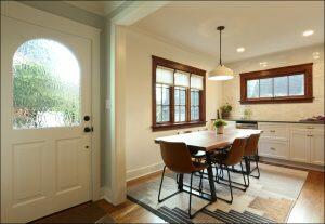 A well-lit dining area with a wooden table, four brown chairs, a hanging light fixture, and windows. The entry door has a textured glass panel. A well-lit dining area with a wooden table, four brown chairs, a hanging light fixture, and windows. The entry door has a textured glass panel.