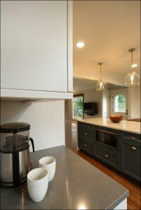 Modern kitchen with gray countertops, a coffee maker, and two white mugs. An island with a microwave, fruit bowl, and pendant lights is visible in the background. Modern kitchen with gray countertops, a coffee maker, and two white mugs. An island with a microwave, fruit bowl, and pendant lights is visible in the background.