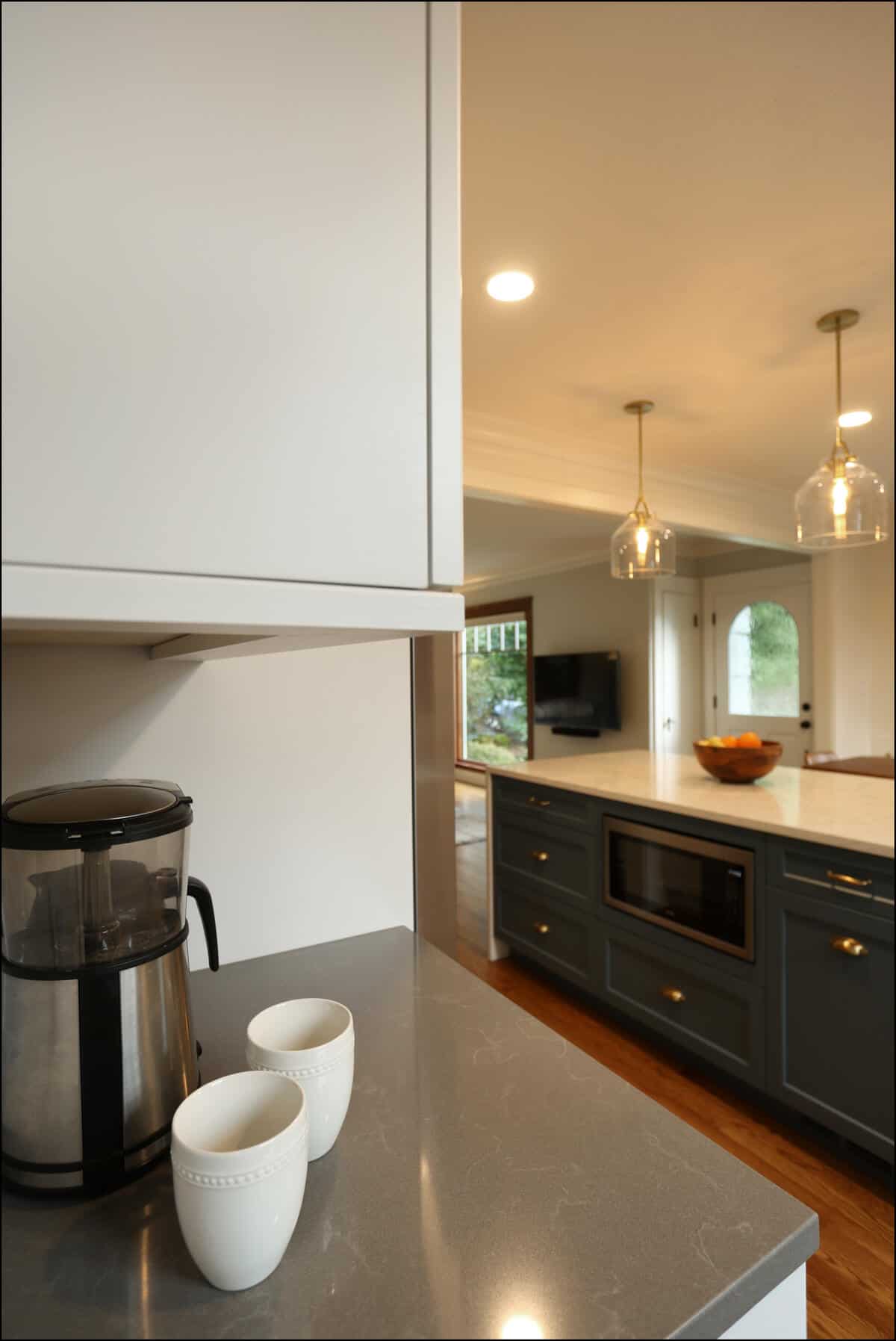 Modern kitchen with gray countertops, a coffee maker, and two white mugs. An island with a microwave, fruit bowl, and pendant lights is visible in the background. Modern kitchen with gray countertops, a coffee maker, and two white mugs. An island with a microwave, fruit bowl, and pendant lights is visible in the background.