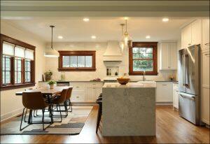 A modern kitchen with white cabinets, marble countertops, a central island, stainless steel appliances, wooden floor, and a dining area with a table and brown chairs adjacent to a large window. A modern kitchen with white cabinets, marble countertops, a central island, stainless steel appliances, wooden floor, and a dining area with a table and brown chairs adjacent to a large window.