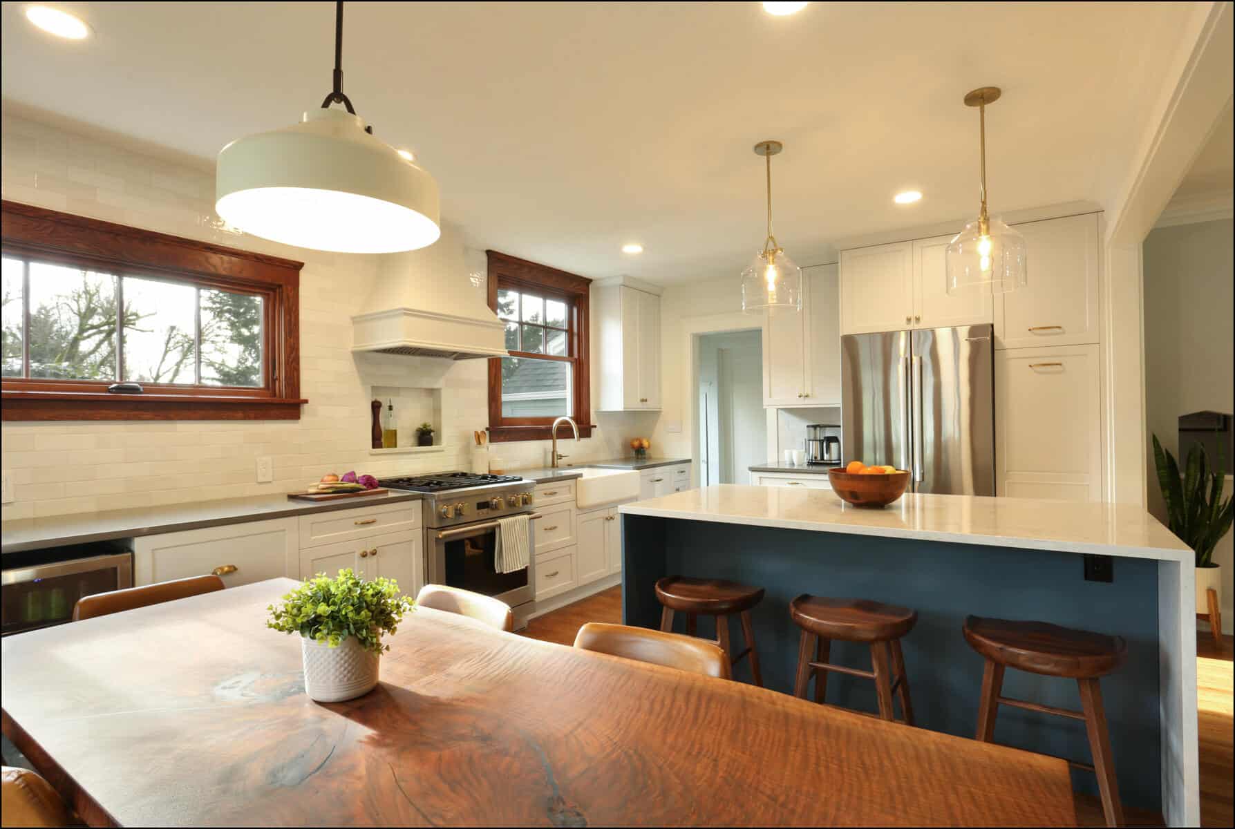 Modern kitchen with wooden dining table, potted plant centerpiece, stainless steel appliances, white cabinets, and island with bar stools and pendant lights. Modern kitchen with wooden dining table, potted plant centerpiece, stainless steel appliances, white cabinets, and island with bar stools and pendant lights.