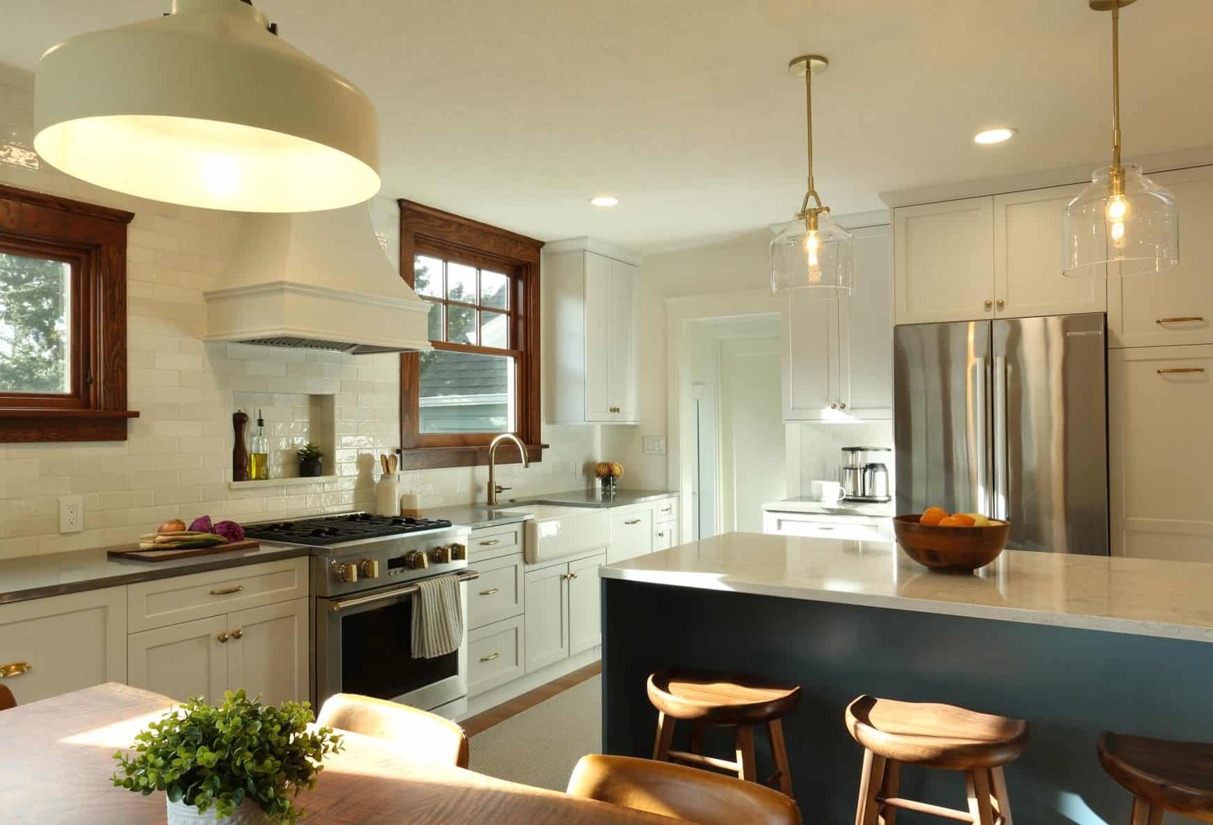 A modern kitchen with white cabinets, stainless steel appliances, an island with wooden stools, pendant lighting, and a bowl of fruit on the countertop. A small plant decorates the dining table. A modern kitchen with white cabinets, stainless steel appliances, an island with wooden stools, pendant lighting, and a bowl of fruit on the countertop. A small plant decorates the dining table.