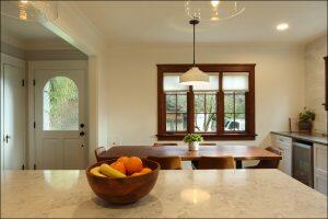 A kitchen interior with a marble countertop, wooden bowl of fruit, dining table, chairs, potted plant, window with blinds, pendant light, door with frosted glass, and a view of trees outside. A kitchen interior with a marble countertop, wooden bowl of fruit, dining table, chairs, potted plant, window with blinds, pendant light, door with frosted glass, and a view of trees outside.