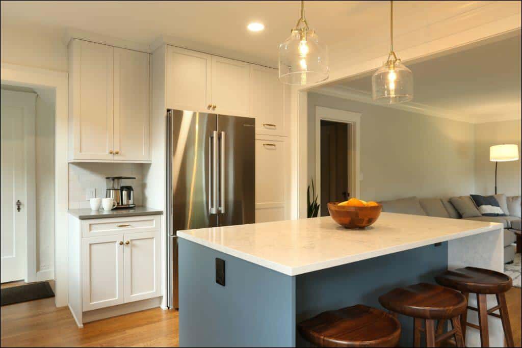 A modern kitchen features a stainless steel refrigerator, white cabinets, a white countertop island with a wooden bowl of oranges, and three wooden stools. Pendant lights hang above the island. A modern kitchen features a stainless steel refrigerator, white cabinets, a white countertop island with a wooden bowl of oranges, and three wooden stools. Pendant lights hang above the island.