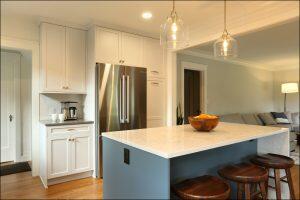 A modern kitchen features a stainless steel refrigerator, white cabinets, a white countertop island with a wooden bowl of oranges, and three wooden stools. Pendant lights hang above the island. A modern kitchen features a stainless steel refrigerator, white cabinets, a white countertop island with a wooden bowl of oranges, and three wooden stools. Pendant lights hang above the island.