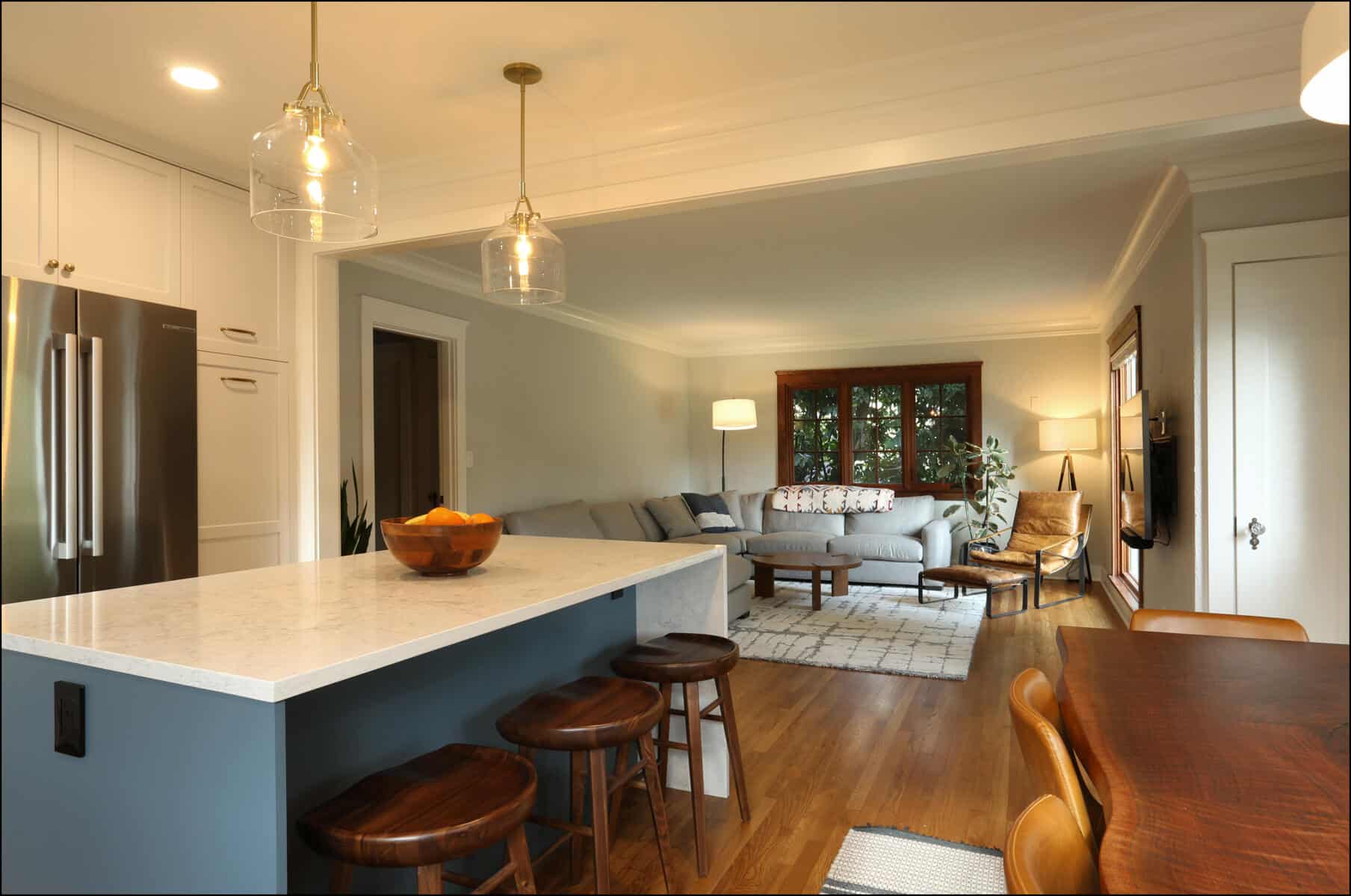 Open-concept kitchen and living room with a marble countertop island, wooden stools, modern light fixtures, a gray sectional sofa, coffee table, and a TV mounted on the wall. Open-concept kitchen and living room with a marble countertop island, wooden stools, modern light fixtures, a gray sectional sofa, coffee table, and a TV mounted on the wall.