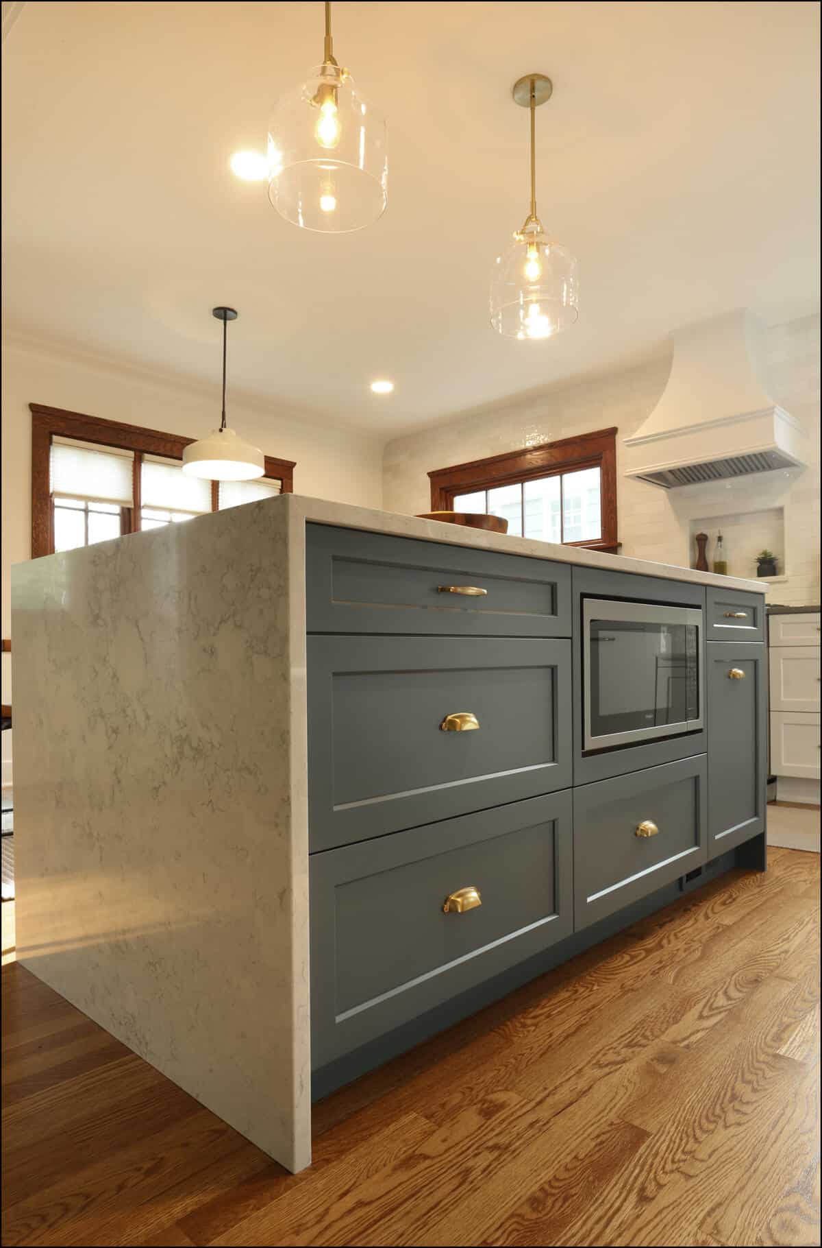 A modern kitchen with a large, marble-top island featuring teal cabinetry, brass handles, and built-in microwave. The room has hardwood floors, pendant lights, and natural light from surrounding windows. A modern kitchen with a large, marble-top island featuring teal cabinetry, brass handles, and built-in microwave. The room has hardwood floors, pendant lights, and natural light from surrounding windows.
