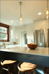 Modern kitchen with white cabinets, stainless steel refrigerator, and pendant lights. A wooden bowl with fruit sits on a marble island countertop, accompanied by wooden bar stools. Modern kitchen with white cabinets, stainless steel refrigerator, and pendant lights. A wooden bowl with fruit sits on a marble island countertop, accompanied by wooden bar stools.