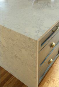 Close-up of kitchen island corner with a marble countertop and blue-grey drawers. The lower half of the image shows a wooden floor. Close-up of kitchen island corner with a marble countertop and blue-grey drawers. The lower half of the image shows a wooden floor.