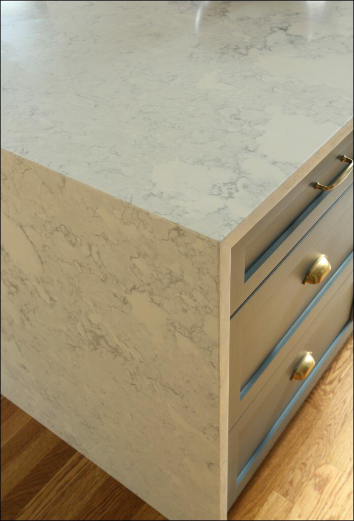 Close-up of kitchen island corner with a marble countertop and blue-grey drawers. The lower half of the image shows a wooden floor. Close-up of kitchen island corner with a marble countertop and blue-grey drawers. The lower half of the image shows a wooden floor.