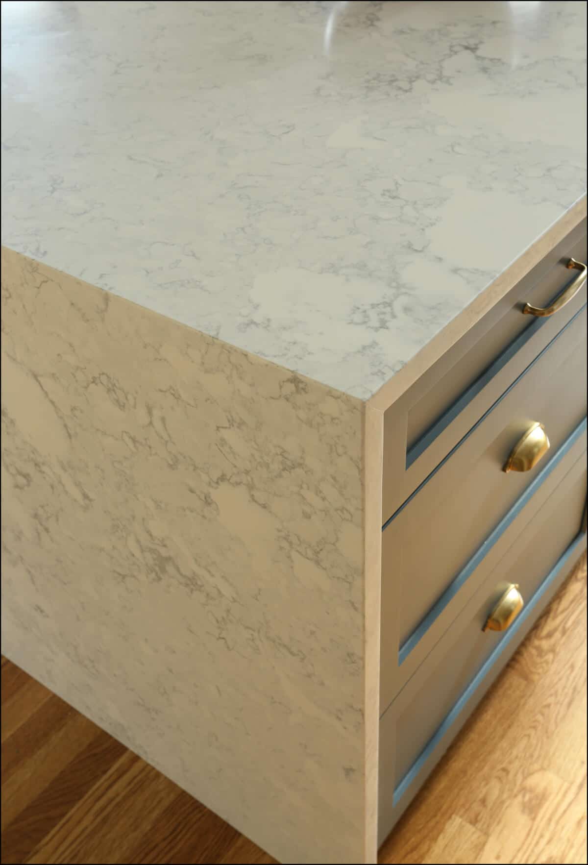 Close-up of kitchen island corner with a marble countertop and blue-grey drawers. The lower half of the image shows a wooden floor. Close-up of kitchen island corner with a marble countertop and blue-grey drawers. The lower half of the image shows a wooden floor.