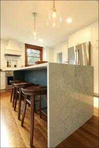 A modern kitchen with a marble island, wooden stools, stainless steel appliances, pendant lights, wooden flooring, and white cabinets. A modern kitchen with a marble island, wooden stools, stainless steel appliances, pendant lights, wooden flooring, and white cabinets.