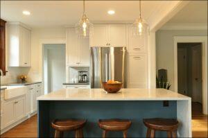 A modern kitchen features a central island with a bowl of fruit, three wooden stools, pendant lights, stainless steel refrigerator, white cabinets, and wooden flooring. A modern kitchen features a central island with a bowl of fruit, three wooden stools, pendant lights, stainless steel refrigerator, white cabinets, and wooden flooring.