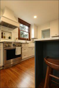 Modern kitchen with white cabinetry, stainless steel stove, white range hood, farmhouse sink, and wooden floors. A striped towel hangs on the oven handle, and a wooden stool is placed at the counter. Modern kitchen with white cabinetry, stainless steel stove, white range hood, farmhouse sink, and wooden floors. A striped towel hangs on the oven handle, and a wooden stool is placed at the counter.