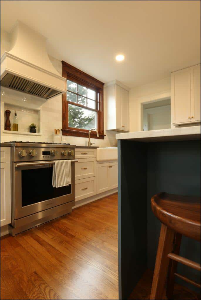 Modern kitchen with white cabinetry, stainless steel stove, white range hood, farmhouse sink, and wooden floors. A striped towel hangs on the oven handle, and a wooden stool is placed at the counter. Modern kitchen with white cabinetry, stainless steel stove, white range hood, farmhouse sink, and wooden floors. A striped towel hangs on the oven handle, and a wooden stool is placed at the counter.