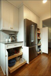 Kitchen with wooden floor, white cabinets, a stainless steel refrigerator, and multiple storage drawers and shelves, including a pull-out pantry beside the fridge and an open cabinet with kitchenware. Kitchen with wooden floor, white cabinets, a stainless steel refrigerator, and multiple storage drawers and shelves, including a pull-out pantry beside the fridge and an open cabinet with kitchenware.