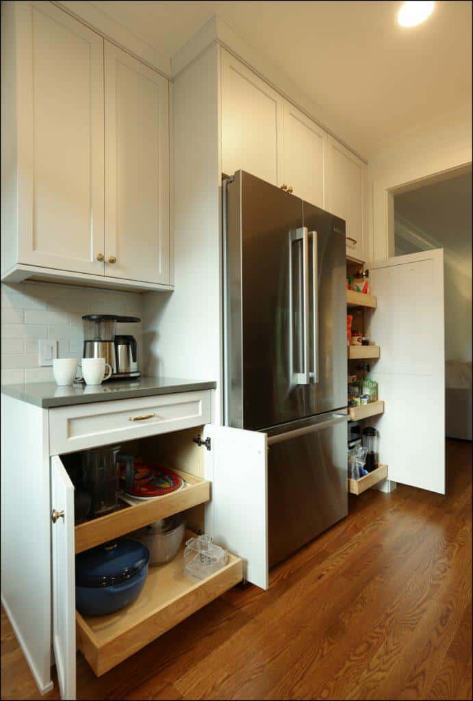 Kitchen with wooden floor, white cabinets, a stainless steel refrigerator, and multiple storage drawers and shelves, including a pull-out pantry beside the fridge and an open cabinet with kitchenware. Kitchen with wooden floor, white cabinets, a stainless steel refrigerator, and multiple storage drawers and shelves, including a pull-out pantry beside the fridge and an open cabinet with kitchenware.