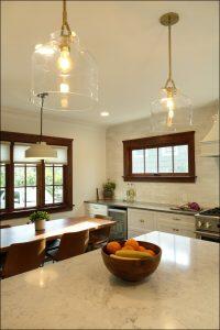A modern kitchen with white cabinets, marble countertop, wooden accents, pendant lights, and a bowl of fruit on the island. A dining area with a wooden table and brown chairs is in the background. A modern kitchen with white cabinets, marble countertop, wooden accents, pendant lights, and a bowl of fruit on the island. A dining area with a wooden table and brown chairs is in the background.