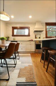 A modern kitchen features white cabinets, a stainless steel stove, and a white range hood. A dining table with brown chairs and a rug is adjacent to the kitchen island with wooden stools. A modern kitchen features white cabinets, a stainless steel stove, and a white range hood. A dining table with brown chairs and a rug is adjacent to the kitchen island with wooden stools.