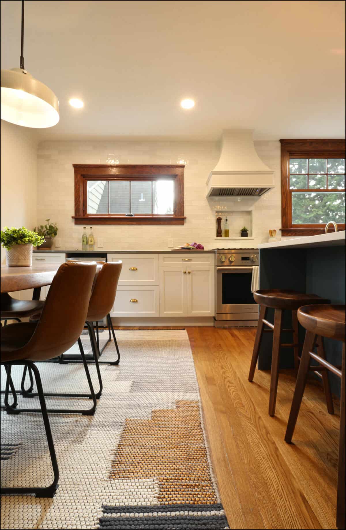 A modern kitchen features white cabinets, a stainless steel stove, and a white range hood. A dining table with brown chairs and a rug is adjacent to the kitchen island with wooden stools. A modern kitchen features white cabinets, a stainless steel stove, and a white range hood. A dining table with brown chairs and a rug is adjacent to the kitchen island with wooden stools.
