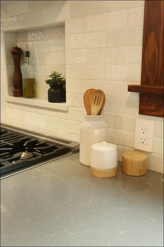 A modern kitchen countertop with a stove, olive oil, and vinegar bottles, a potted plant, a container holding wooden utensils, and a ceramic jar with a wooden lid in a white-tiled backsplash setting. A modern kitchen countertop with a stove, olive oil, and vinegar bottles, a potted plant, a container holding wooden utensils, and a ceramic jar with a wooden lid in a white-tiled backsplash setting.