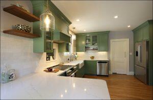 A modern kitchen with green cabinets, a white marble countertop, stainless steel appliances, wooden shelves, and a pendant light above the island. The floor is wooden, and the walls are light gray.