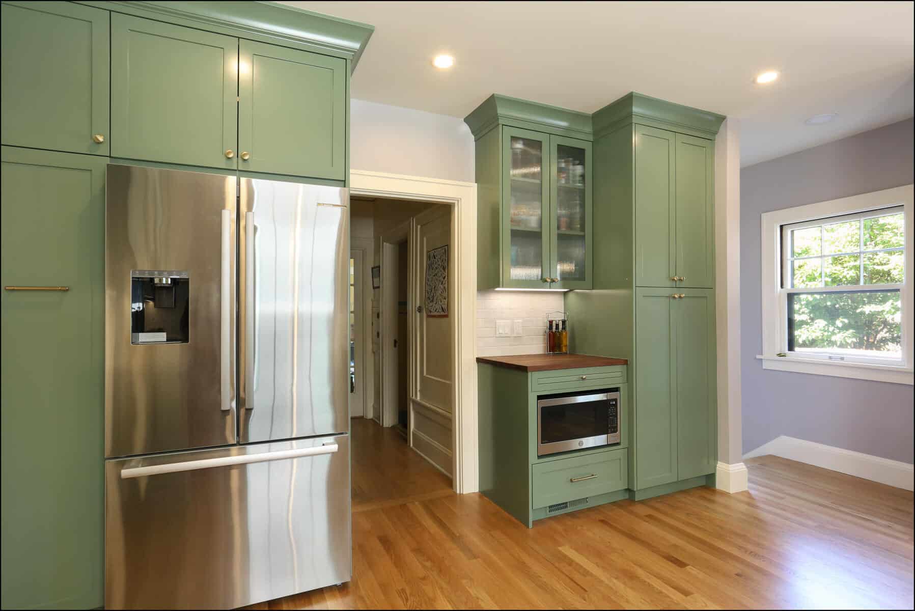 Modern kitchen with green cabinets, stainless steel refrigerator, built-in microwave, and hardwood flooring. White walls and a window providing natural light. Modern kitchen with green cabinets, stainless steel refrigerator, built-in microwave, and hardwood flooring. White walls and a window providing natural light.
