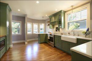 Modern kitchen with green cabinetry, hardwood floors, white countertops, a farmhouse sink, stainless steel appliances, and a bay window with seating area.